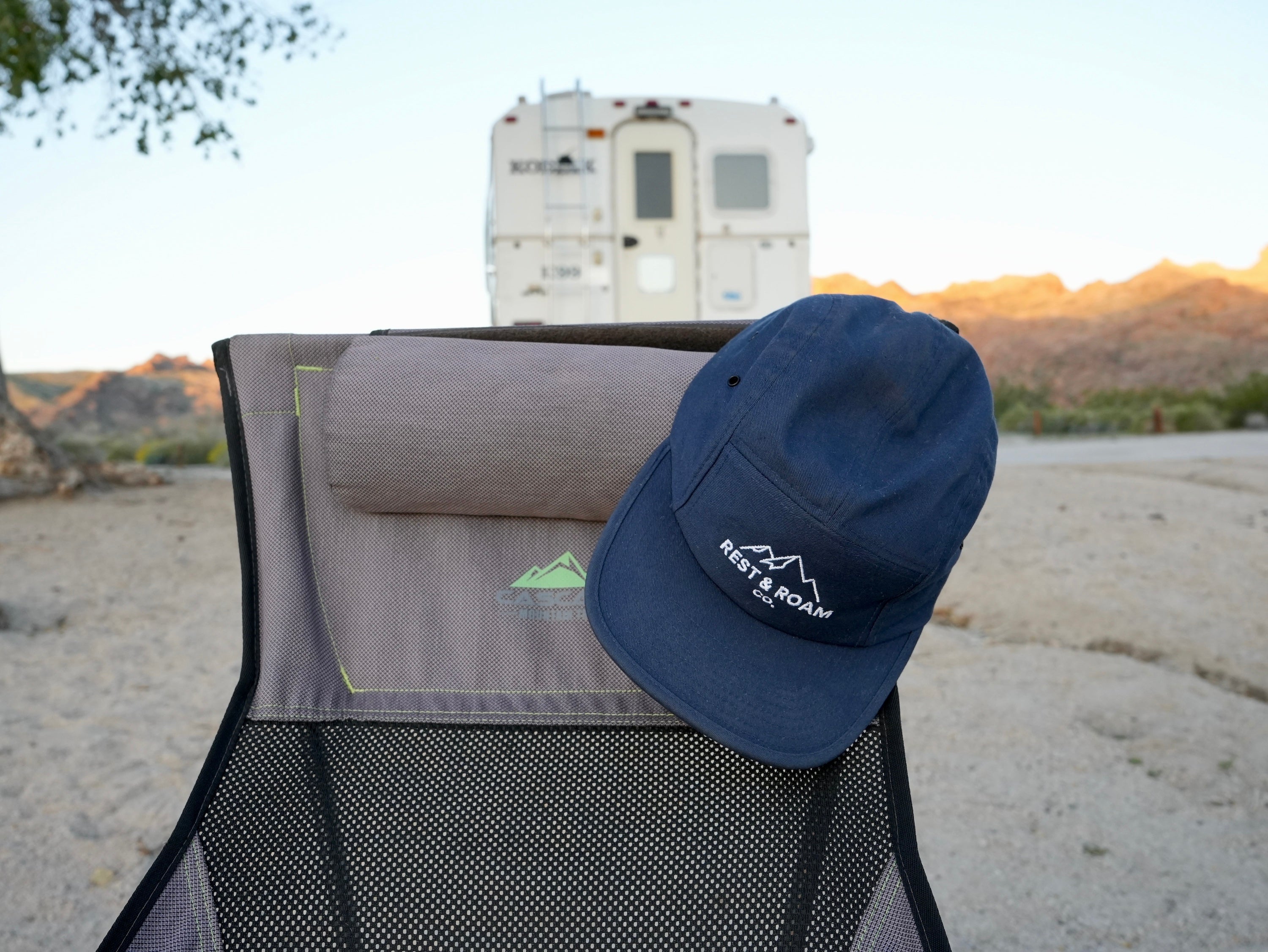 Blue 5-panel camp cap with Rest & Roam Co. logo on top of a camping chair with a scenic background and truck camper