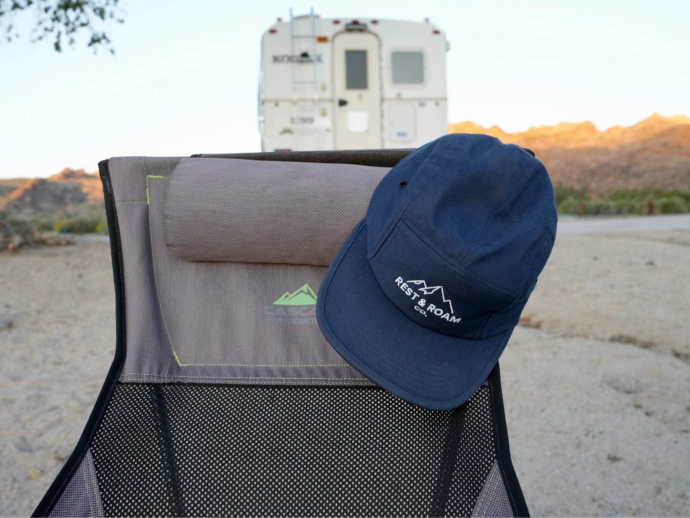 Blue 5-panel camp cap with Rest & Roam Co. logo on top of a camping chair with a scenic background and truck camper