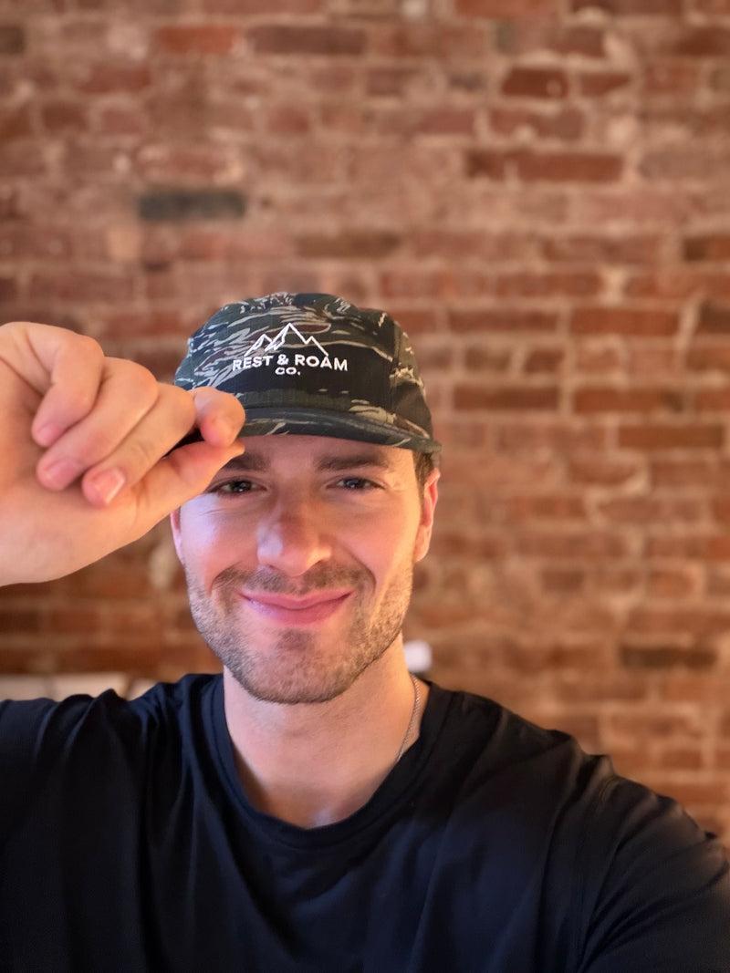 Man holding a camo 5-panel camp hat with Rest & Roam Co. logo in front of a brick wall