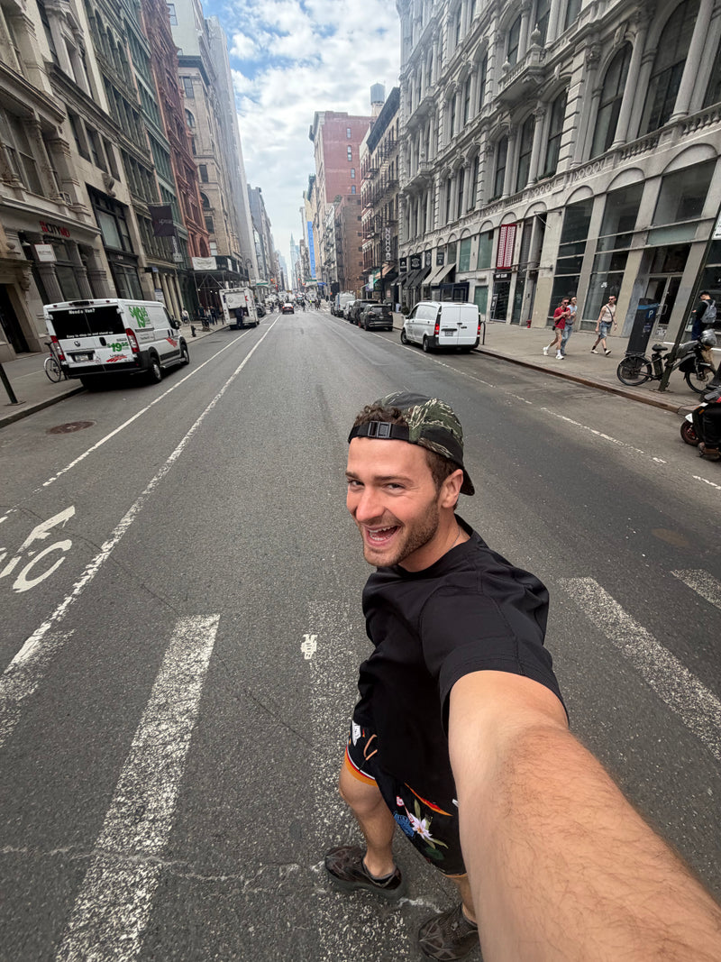 Man taking a selfie on a city street with buildings and vehicles in the background wearing a camo 5-panel camp hat | Rest & Roam Co.