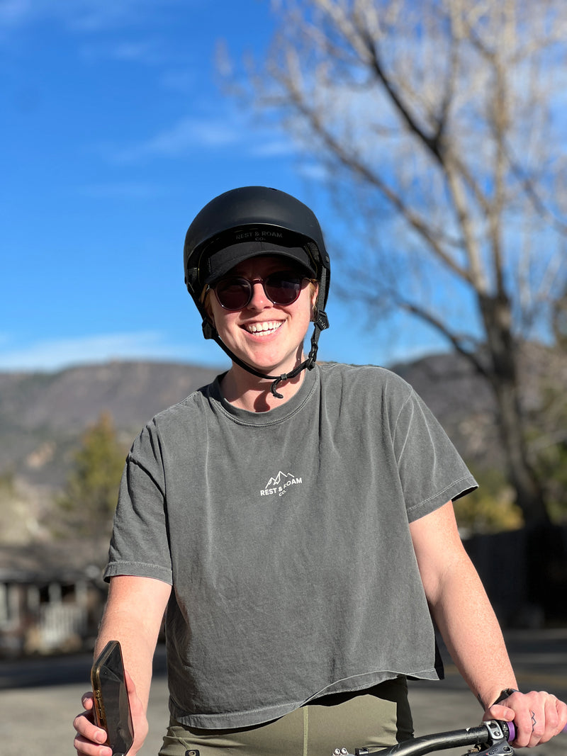 Woman wearing a boxy Tee and helmet, holding a phone, with a scenic background of mountains and trees on a bike ride.