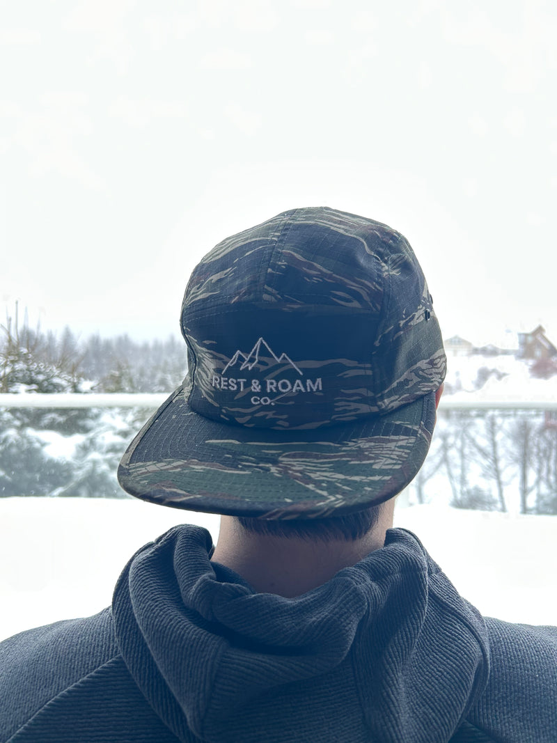 Person wearing a camouflage 5-panel camp hat with Rest & Roam Co. logo in a snowy landscape