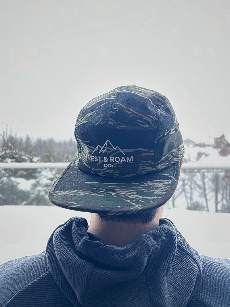 Person wearing a camouflage 5-panel camp hat with Rest & Roam Co. logo in a snowy landscape