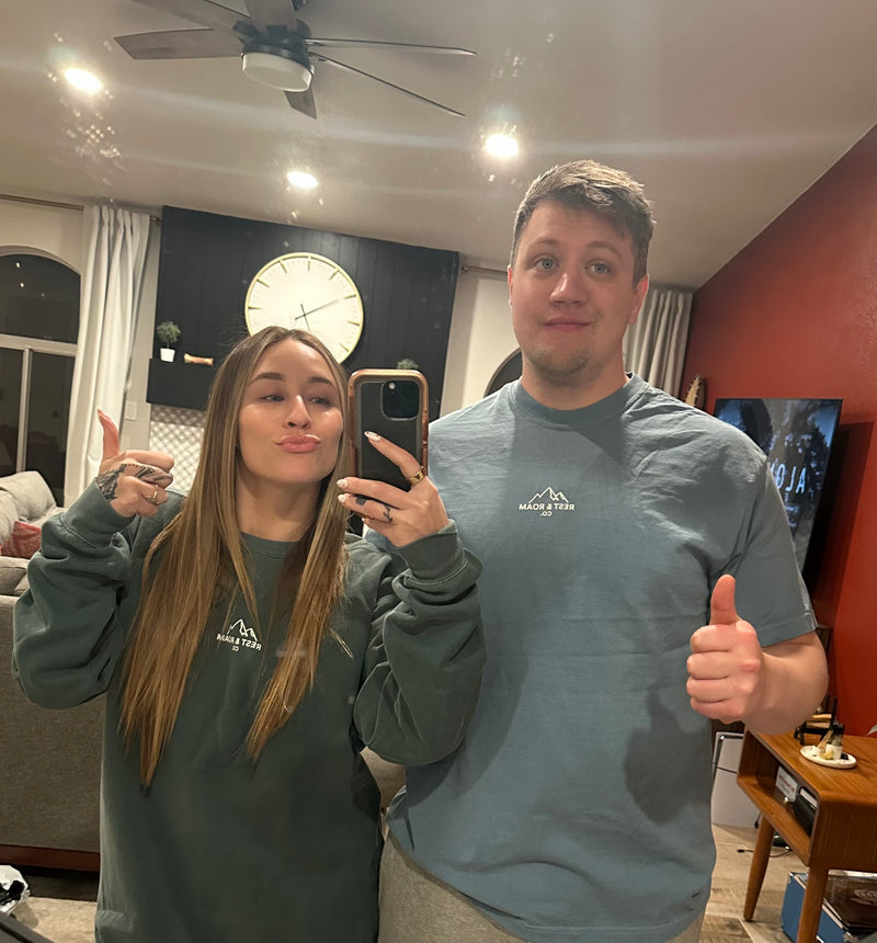 Woman wearing a Rest & Roam Co. crewneck sweatshirt and a man with Rest & Roam Outdoor t-shirt taking a mirror picture in a living room.