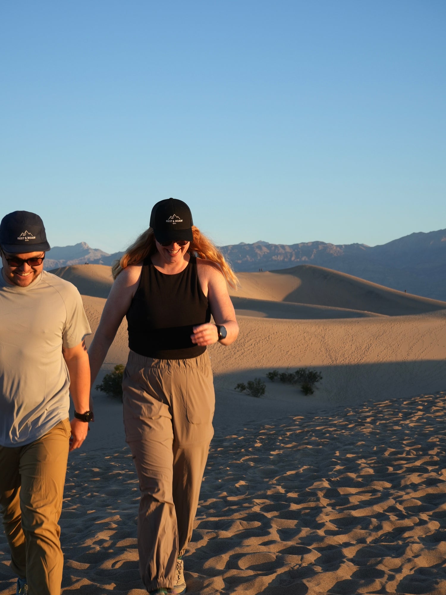Two people wearing a trucker hat and 5-panel camp hat while walking around in a desert landscape at sunrise.