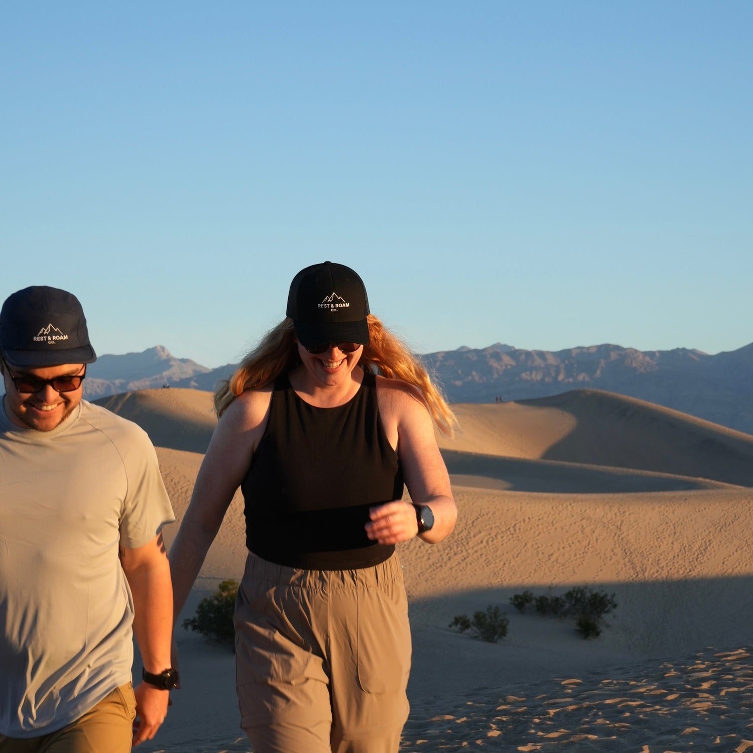 Two people wearing Rest & Roam Co. hats walking through a desert landscape at sunrise. 