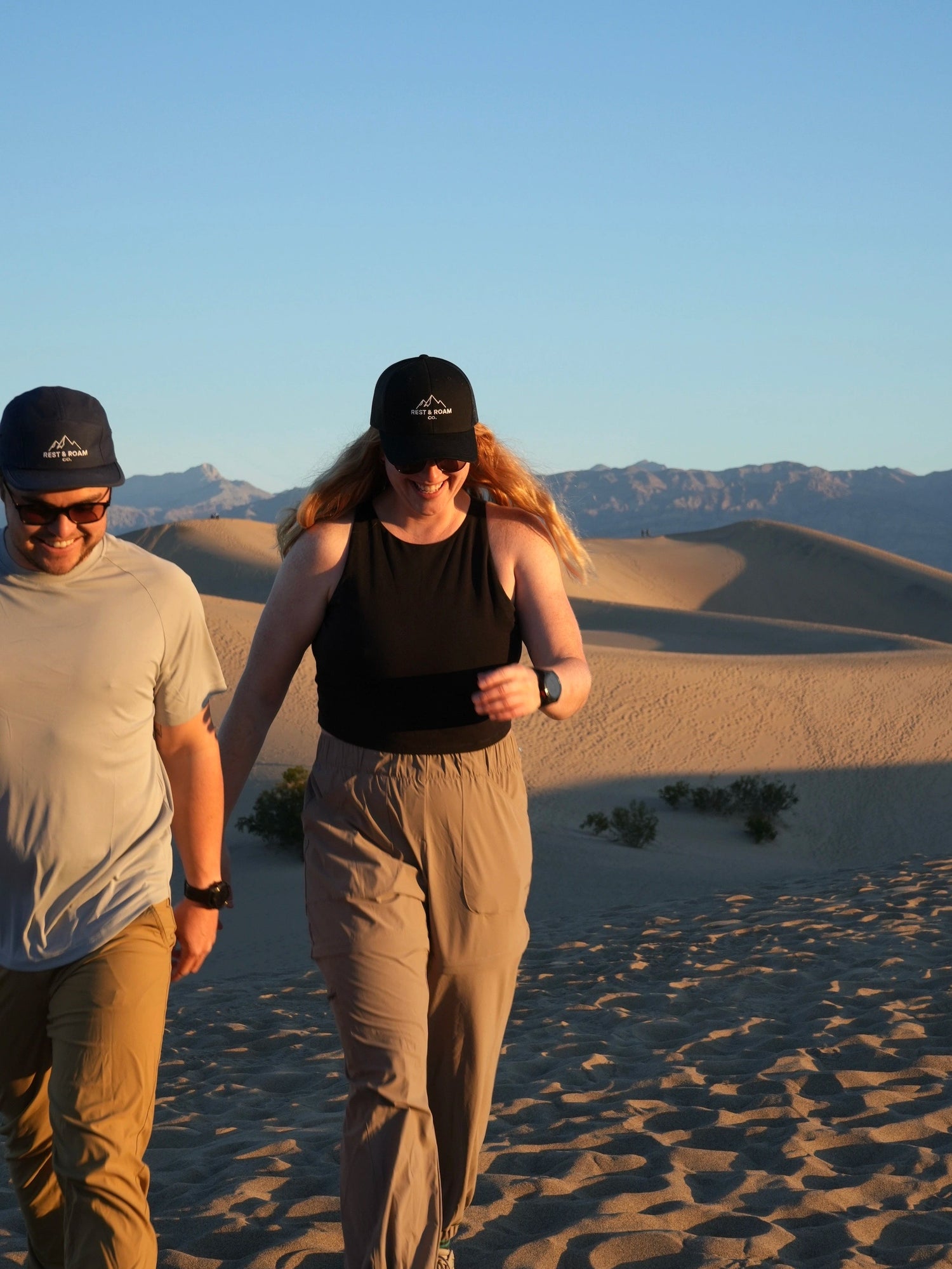 Two people wearing Rest & Roam Co. hats walking through a desert landscape at sunrise.