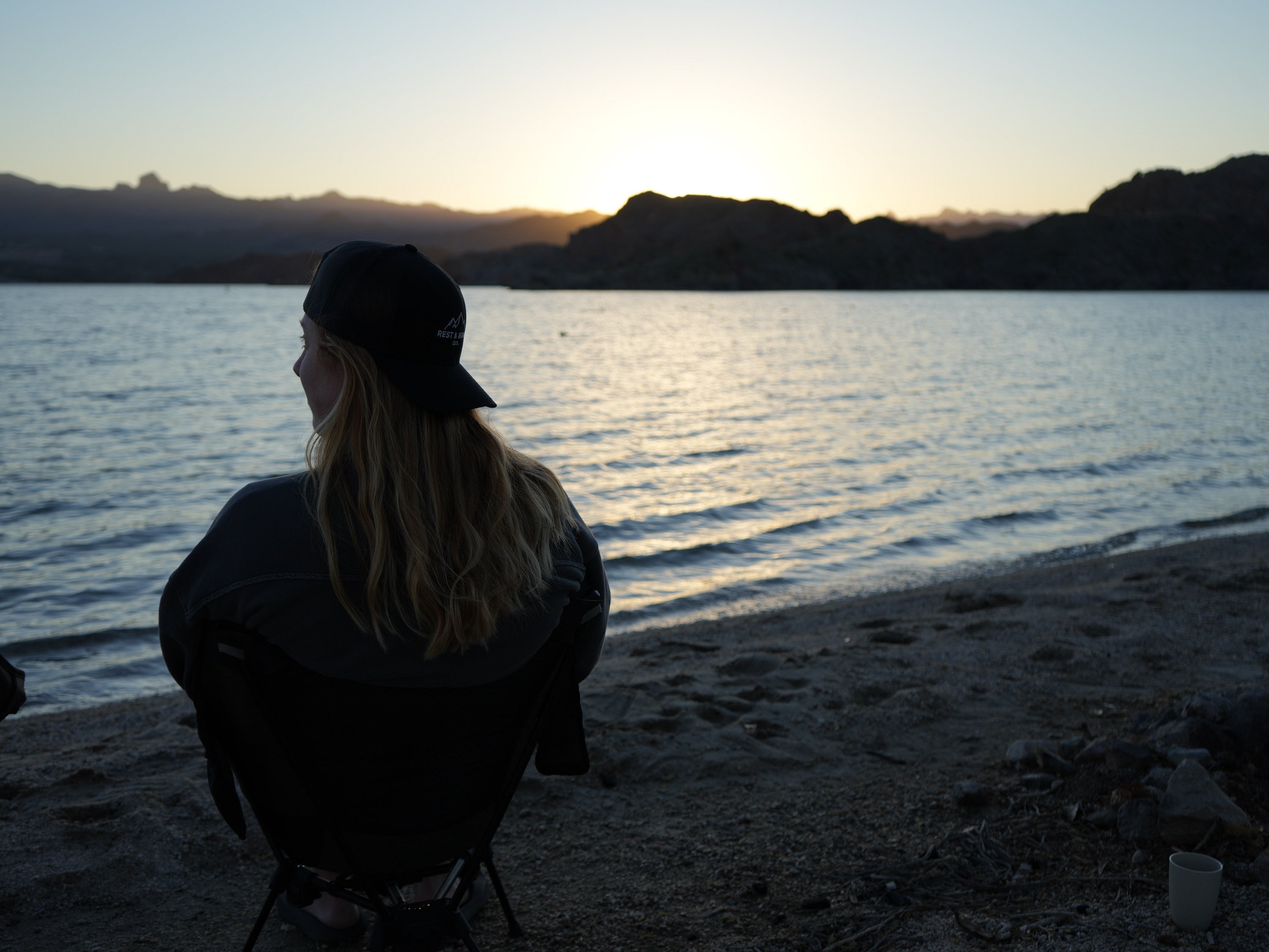 A woman sitting on the beach next to a lake wearing Rest & Roam Co. Trucker Hat at sunrise
