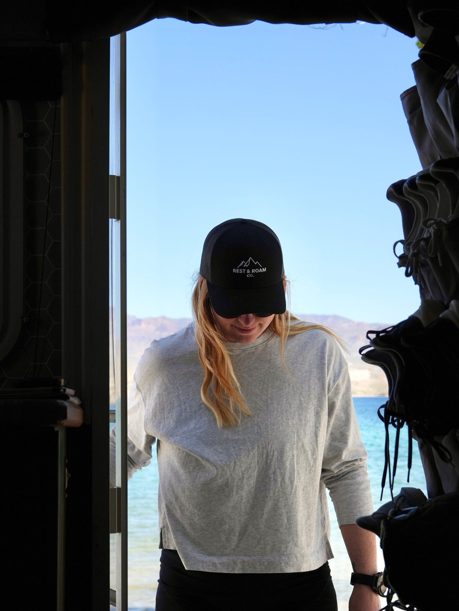 Woman wearing a black trucker cap with a Rest & Roam Co. logo, standing by a door with a scenic view outside near Colorado River
